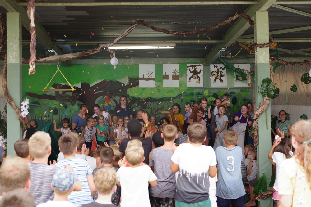Eine Gruppe Kinder vor einem selbstgemalten Dschungel, die Konfetti in die Luft werfen.
